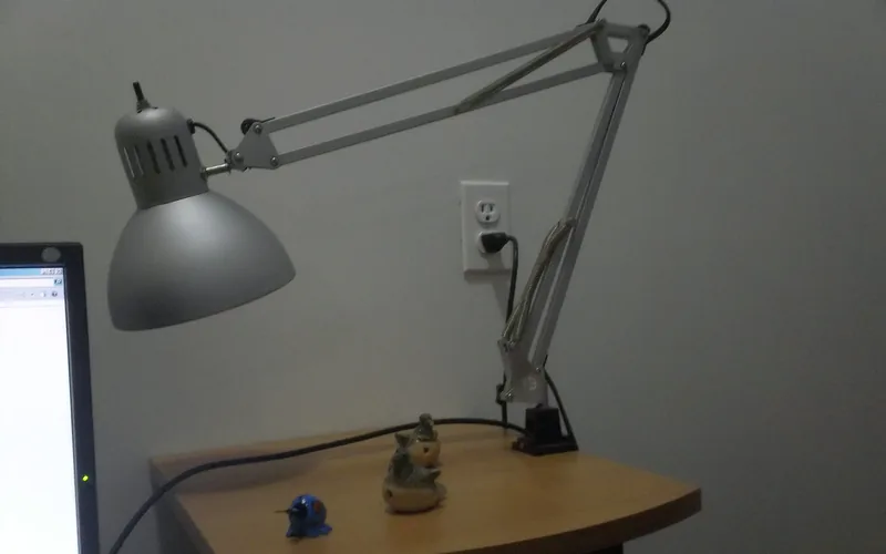 Flexo plateado de muelles sujeto con una mordaza al borde de una mesita, junto a un enchufe de pared y al canto de un monitor