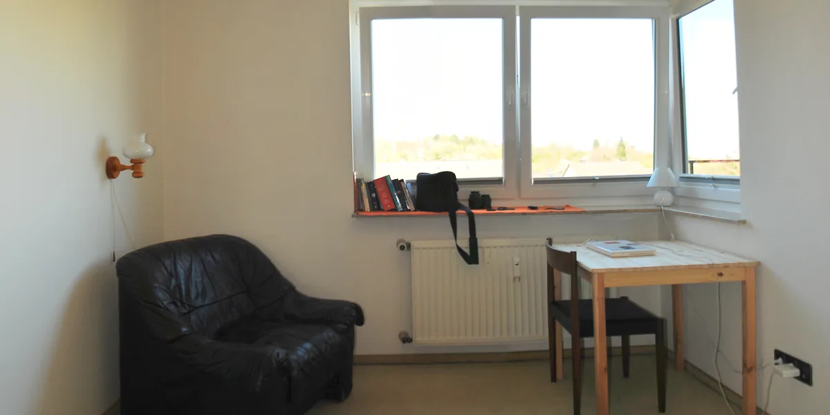 Habitación blanca con una butaca de cuero negro, un radiador bajo una ventana de tres hojas, libros en el alféizar y una mesa de pino con su silla