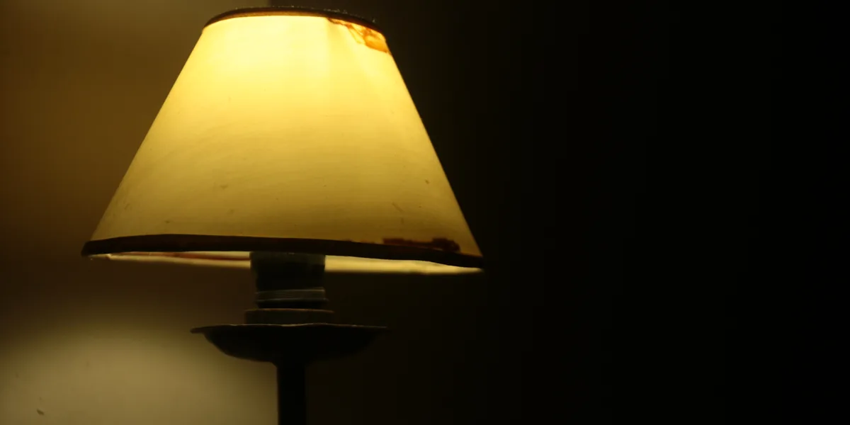 Lámpara de sobremesa con tulipa clara encendida en una habitación oscura; bajo la tulipa asoma el portalámparas